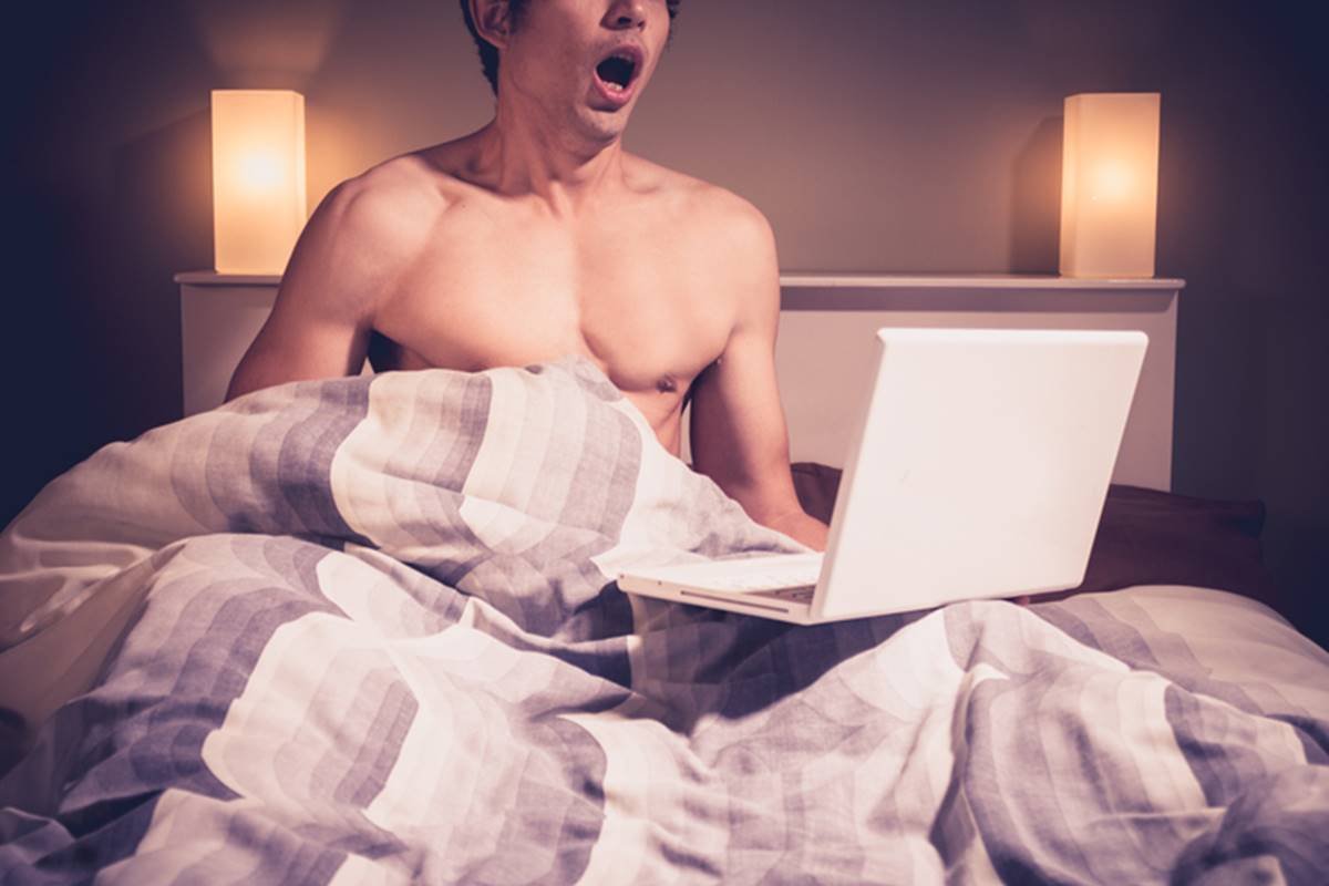 Foto colorida de homem na cama com notebook assistindo pornografia