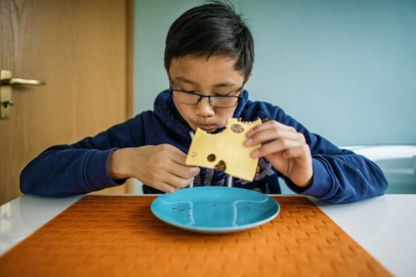 Foto colorida de criança sentada em mesa comendo queijo, bactéria
