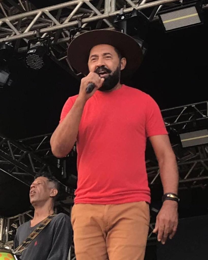 homem de camiseta vermelha, calça caqui e chapeu cantando em palco - metrópoles