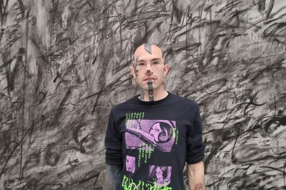 Na foto, homem posa de frente com a blusa preta e mostra o rosto com tatuagens e piercings - Metrópoles