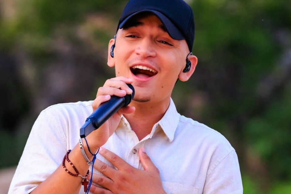 Imagem colorida mostra o cantor João Gomes cantando no microfone. Ele veste uma camisa branca e boné preto - Metrópoles