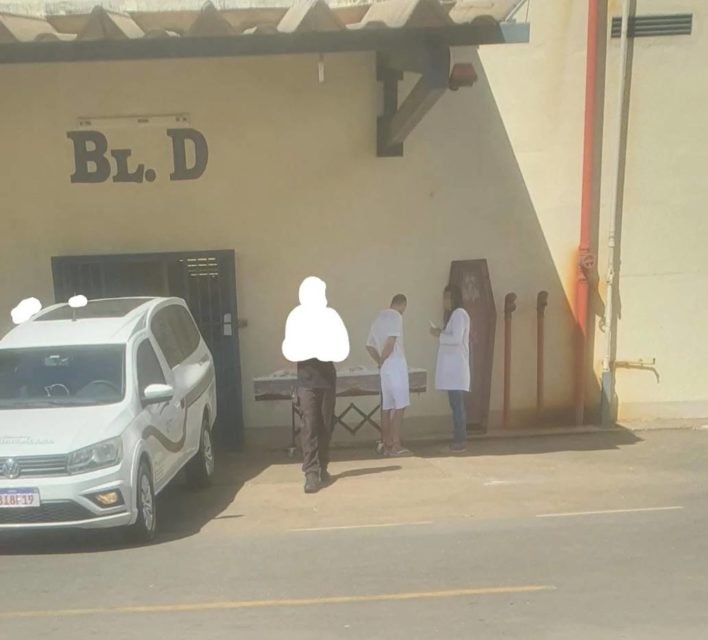 Homem com roupa branca em pé ao lado de caixão e viatura da polícia penal - Metrópoles