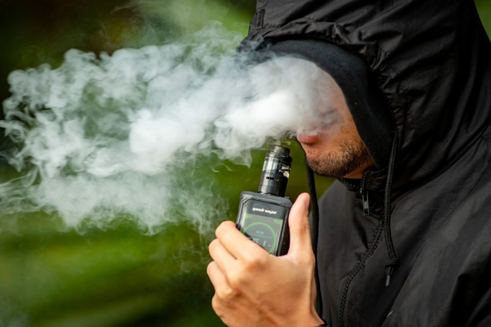 homem branco com casaco preto segurando um cigarro eletrônico
