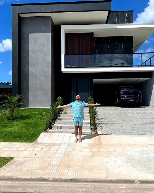 Gil do Vigor de camisa azul de braços abertos na parte externa da mansão