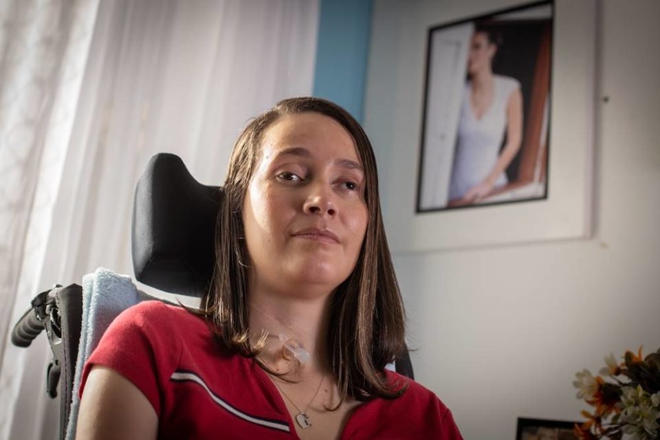Foto colorida de uma mulher branca com cabelos curtos e blusa vermelha. Ela está sentada em uma cadeira de rodas - metrópoles