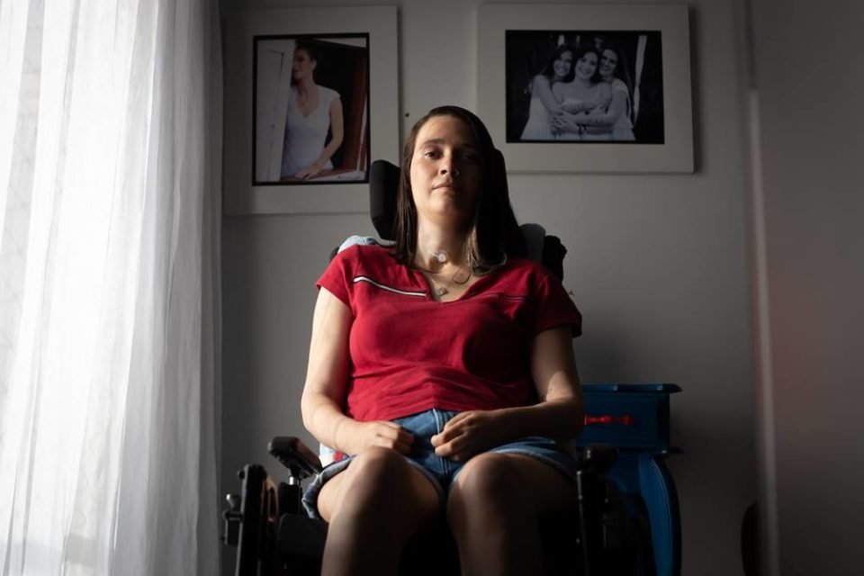 Foto colorida de uma mulher branca com cabelos curtos e blusa vermelha. Ela está sentada em uma cadeira de rodas - metrópoles