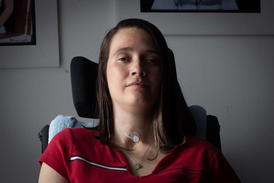 Foto colorida de uma mulher branca com cabelos curtos e blusa vermelha. Ela está sentada em uma cadeira de rodas - metrópoles
