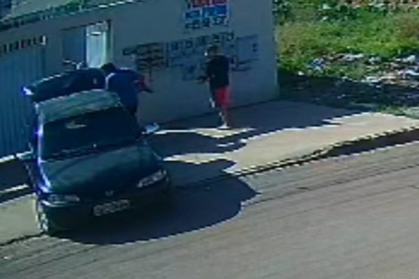 adolescente de bermuda vermelha entra casa carregando uma pipa enquanto dois homens levam uma televisão de 70 polegadas e colocam em um carro preto