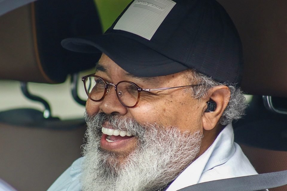 Foto colorida de Jorge Aragão. Ele está com um boné preto e branco, óculos de grau e blusa branca - Metrópoles