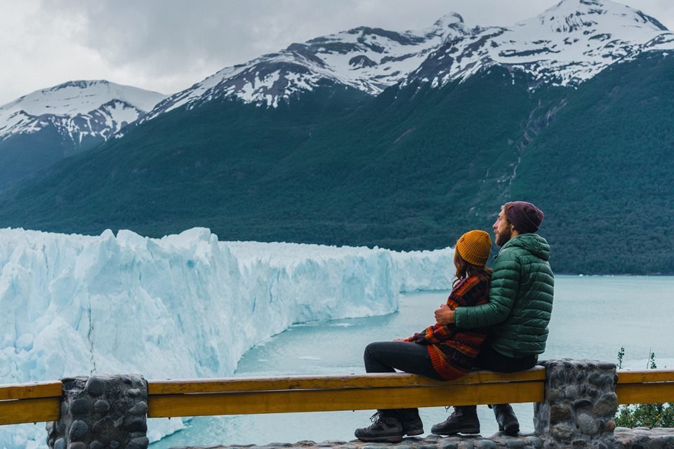 Imagem colorida mostra casal próximo a geleira na cidade de Perito Moreno, na Argentina - Metrópoels