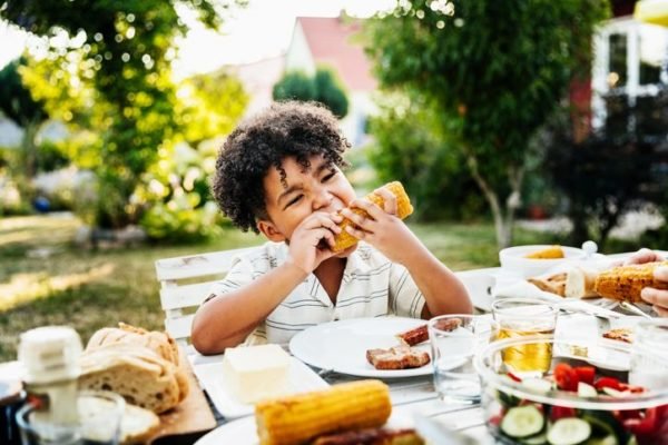 Foto de uma criança negra comendo um milho assado diante de uma bandeja com diversos alimentos