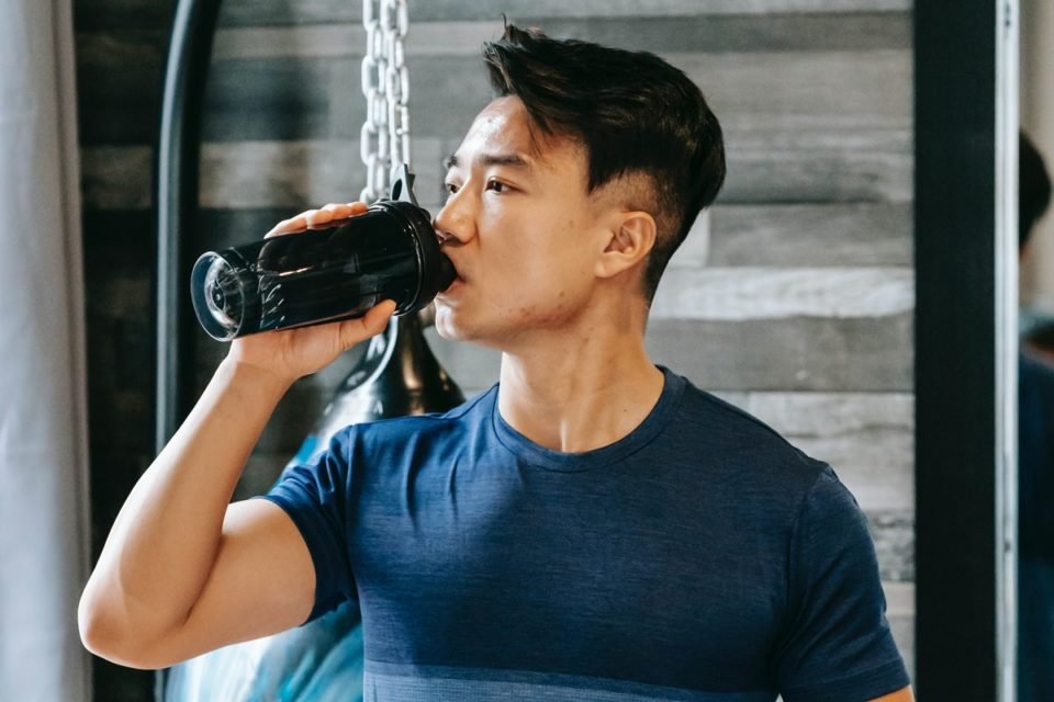 Jovem asiático bebendo shake em garrafinha. Ao fundo, um ambiente de academia - Metrópoles