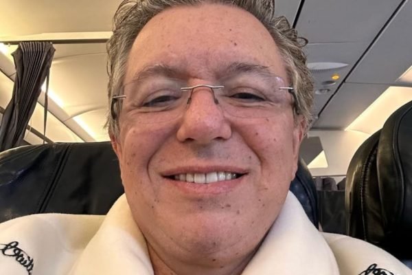 Boninho usa casaco bege e posa dentro de um avião - Metrópoles