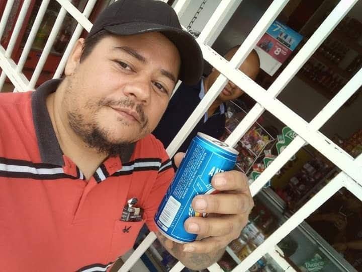 Homem pardo de cabelos negros segura lata de cerveja em frente a portão branco. Ele veste boné e uma camisa polo vermelha com listras pretas