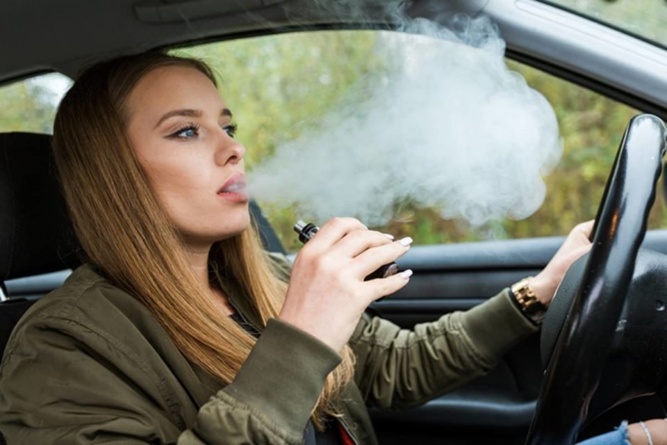 Imagem colorida de jovem dirigindo enquanto fuma tipo de cigarro eletrônico - Metrópoles