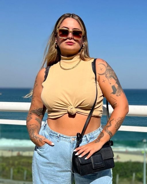 Tatah Fávero está com uma camisa clara, amarrada na barriga, calça jeans e bolsa preta - Metrópoles