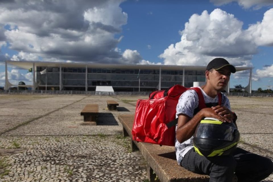 Foto do Documentário brasiliense Da Porta Pra Fora. Um motoboy aparece sentado com o Palácio do Planalto ao fundo - Metrópoles