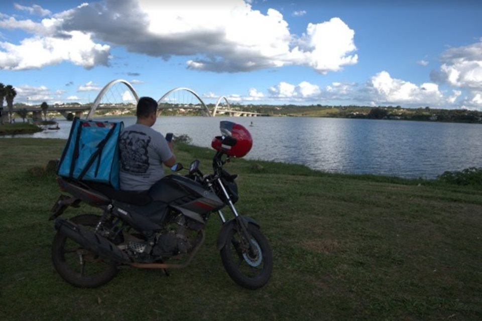 Foto do Documentário brasiliense Da Porta Pra Fora. Motoboy em cima de uma moto na grama com a Ponte JK ao fundo - Metrópoles