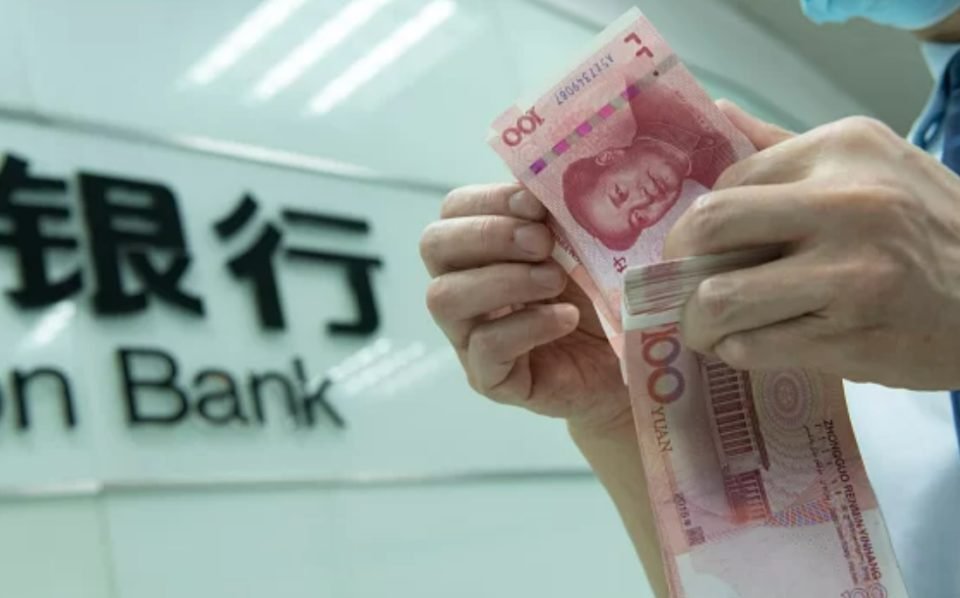 Imagem de notas de dinheiro sendo manuseadas por homem. Ao fundo, logotipo do Banco Central da China