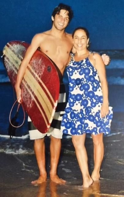 foto colorida de rafael mascarenhas de bermuda segurando uma prancha abraçado em cissa guimarães de vestido azul e branco - metrópoles