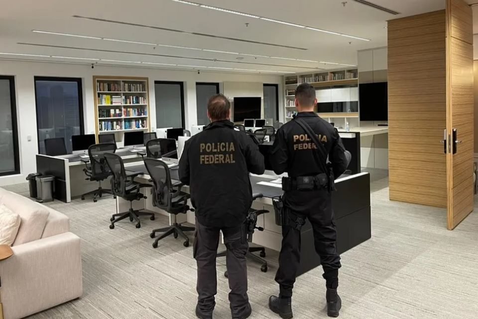 Imagem de dois agentes da Polícia Federal, vestidos com coletes pretos, em operação que investiga a Braiscompany. Eles estão em um escritório. Ao fundo, mesas e cadeiras vazias.