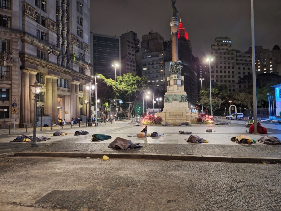 Imagem mostra pessoas dormindo no chão em frente a monumento - Metrópoles