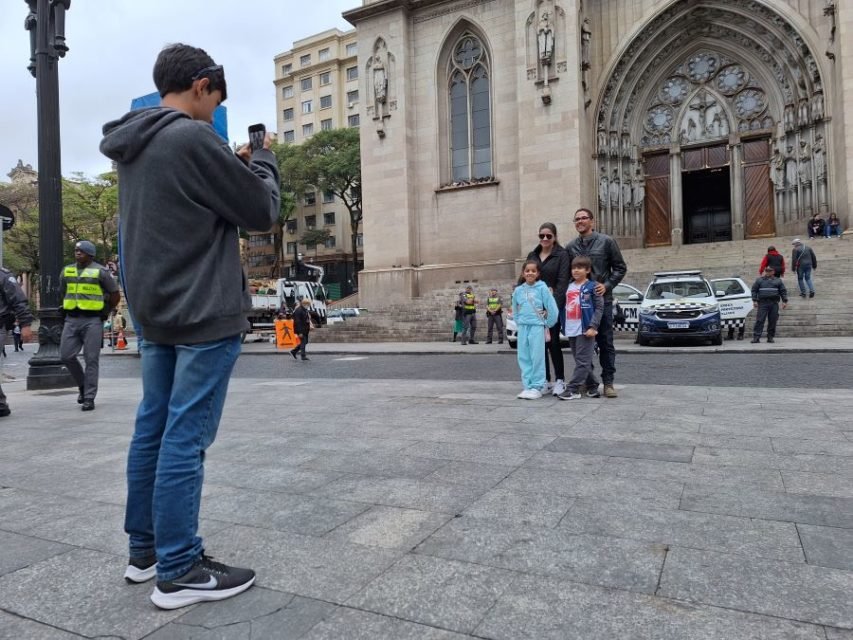Imagem mostra jovem tirando fotos de mulher, homem e duas crianças em frente a igreja
