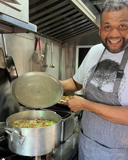 Foto colorida de Wilson Cabral. Ele é negro, tem cabelo preto, usa óculos e está utilizando um avental de cozinheiro. Ele mostra uma panela com comida - Metrópoles