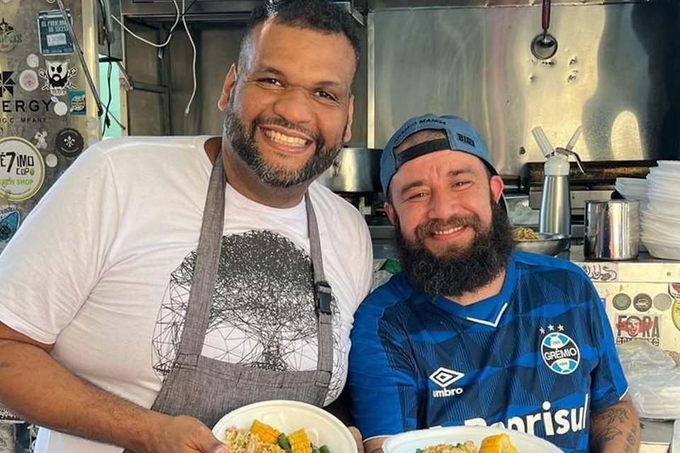 imagem colorida de Hichel Rodrigues com a camisa do grêmio, boné azul, tem barba e cabelo preto. Wilson está de camisa branca, tem barba e cabelo preto. Eles mostram um prato de comida - Metrópoles