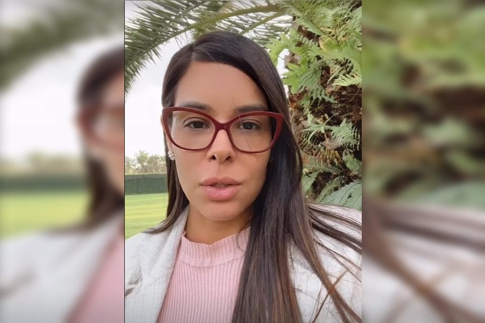 Mulher branca, de cabelos lisos e castanhos, óculos com armação vermelha, olhando para a câmera. Ao fundo, há uma árvore e um gramado - Metrópoles
