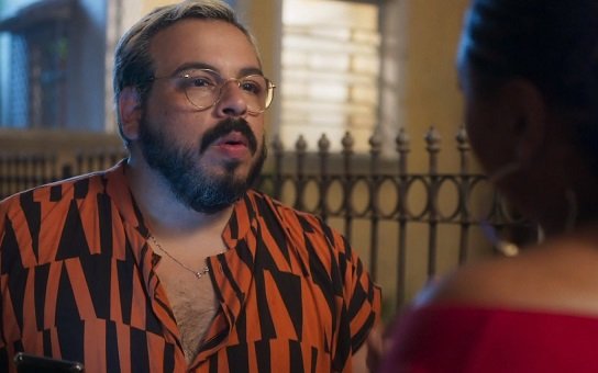 Foto colorida de Luis Lobianco. O ator é branco, usa óculos de grau e está de blusa laranja com preto - Metrópoles