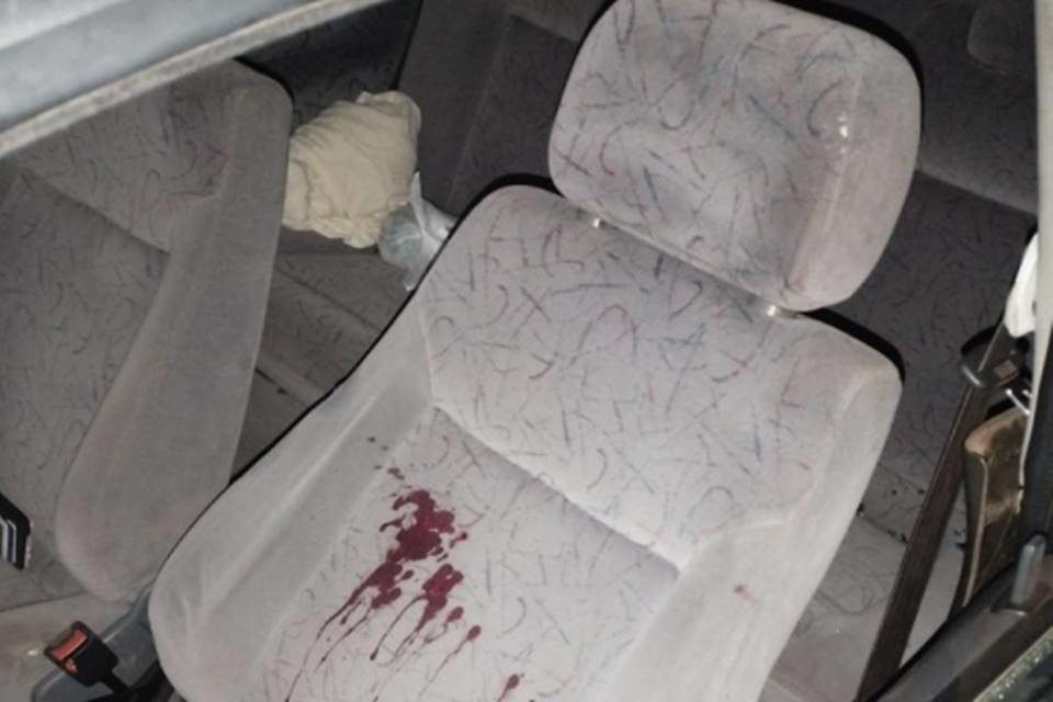 Banco de carro na cor cinza, com mancha de sangue escorrendo; objetos indefinidos deixados ao fundo, no banco traseiro