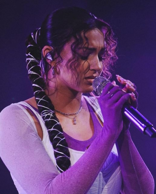 mulher com cachos soltos na frente do rosto e o resto do cabelo preso em trança cumprida cantando de olhos fechados - metrópoles