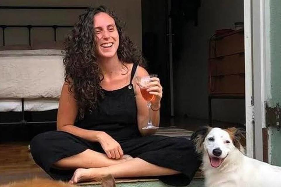 mulher de cabelos cacheados veste macacão preto e está sentada no chão rindo e bebendo vinho ao lado de cachorro - metrópoles