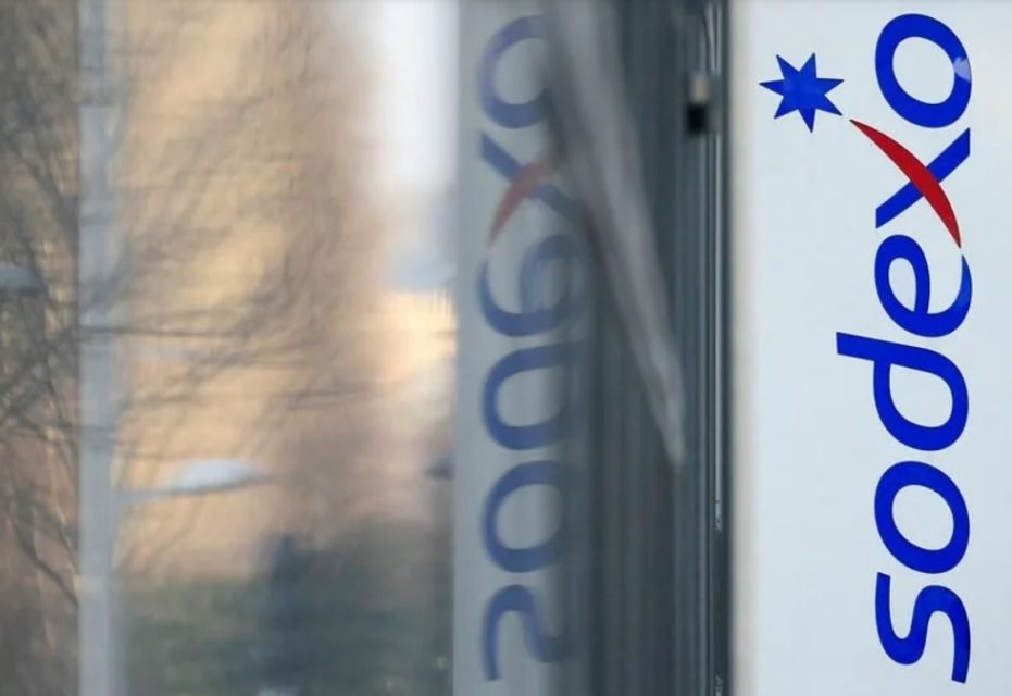 Imagem colorida de logotipo da Sodexo, com letras em azul e vermelho, em uma parede branca. Ao fundo, reflexo da imagem em um espelho - Metrópoles