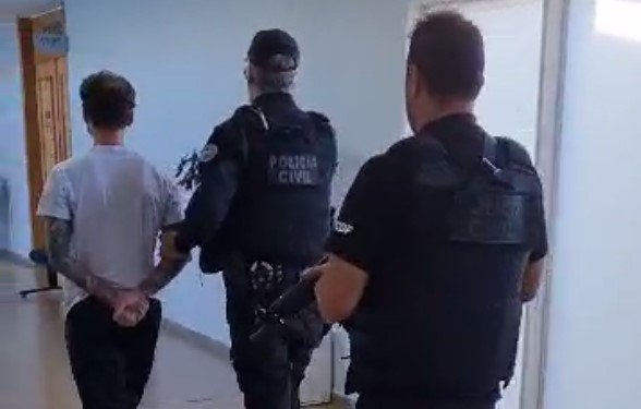 De costas, dois policiais civis com uniformes pretos conduzem homem branco, também de costas, ele usa camiseta branca e calça preta e tem tatuagens no braço