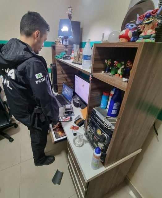 Policial com uniforme preto da PCDF em frente a estante e escrivaninha com computador e diversos outros objetos de item pessoal