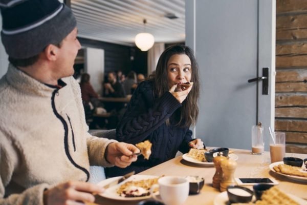 Imagem colorida de homem e mulher sentados em mesa farta comendo muito usando roupas de frio - Metrópoles