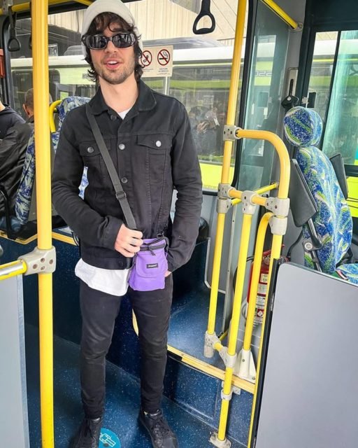 Foto colorida de Fiuk. Ele está no ônibus, com um gorro cinza, roupa preta e bolsa roxa - Metrópoles