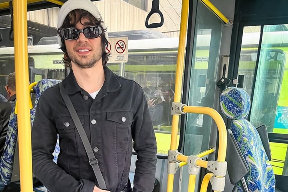 Foto colorida de Fiuk. Ele está no ônibus, com um gorro cinza, roupa preta e bolsa roxa - Metrópoles