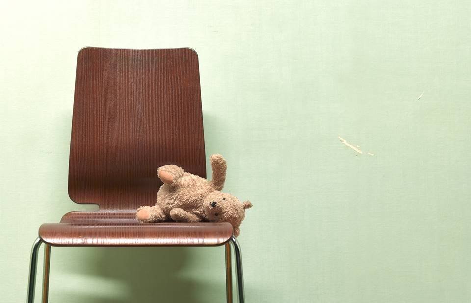 Foto de uma cadeira de madeira com um ursinho de pelúcia caído, com parede verde ao fundo - Metrópoles