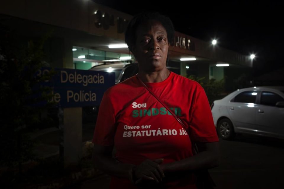 Imagem de mulher negra com camiseta vermelha em frente a delegacia olhando para a câmera - Metrópoles