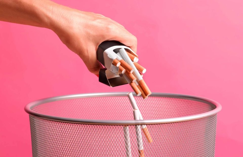 Foto mostra pessoa descartando cigarros em uma lixeira com um fundo monocromático rosa