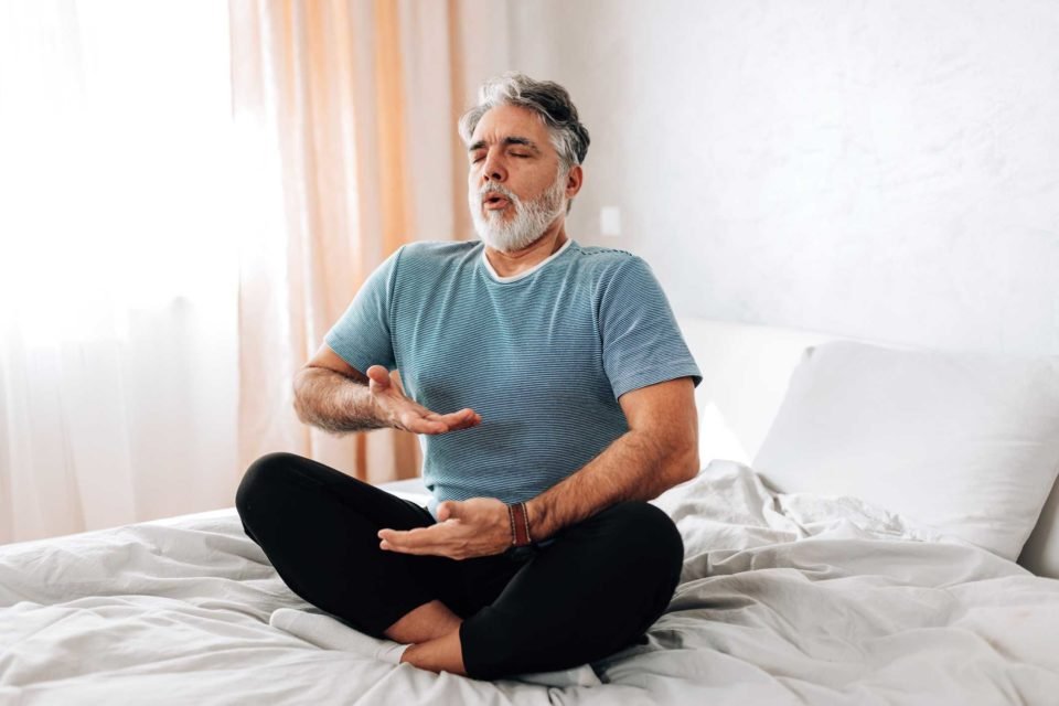 Foto mostra homem idoso em cama praticando yoga e trabalhando a respiração - Alzheimer