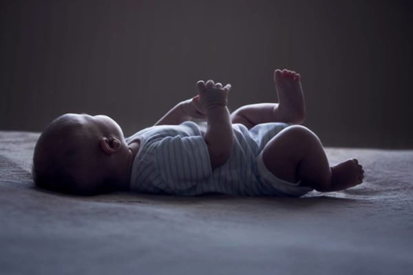 Imagem colorida de bebê contra luz olhando para o lado deitado sozinho - Metrópoles