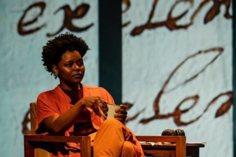 mulher de roupa laranja sentada tomando algo em uma xícara posa séria em cena de peça de teatro - metrópoles