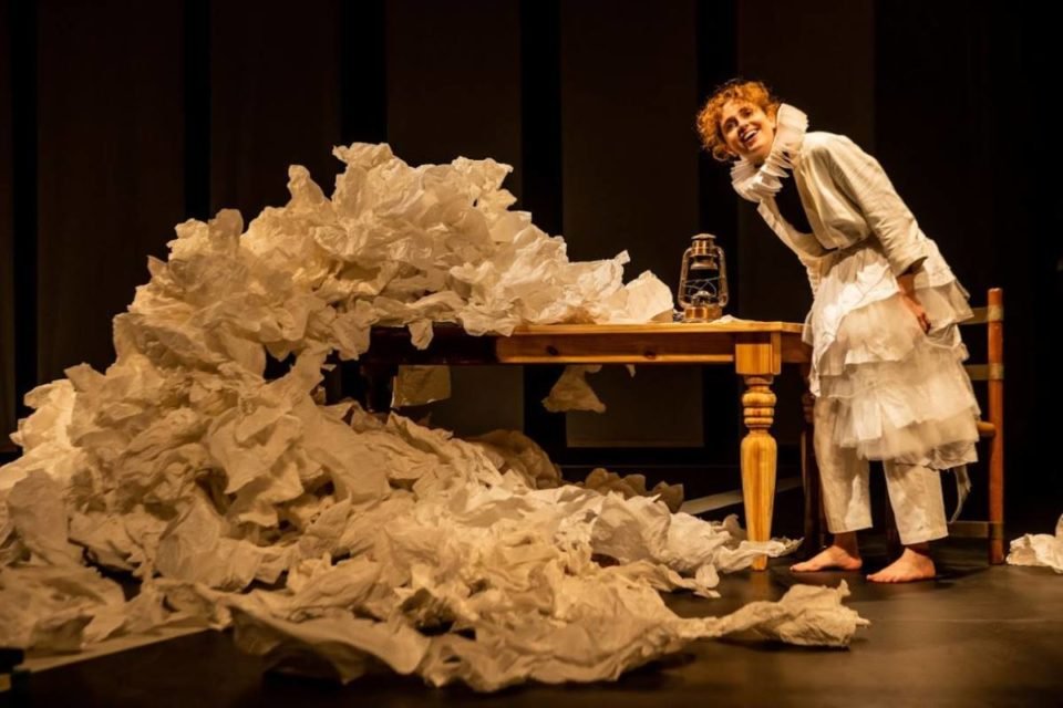 mulher loira com roupas brancas em palco de teatro ao lado de mesa e montanha de papeis - metrópoles
