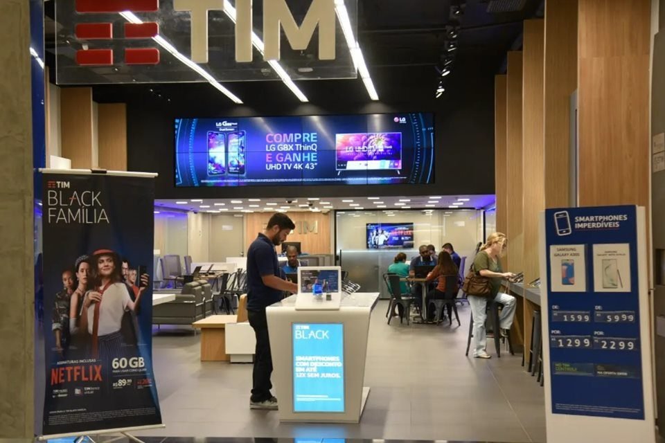 Imagem colorida de entrada de uma loja da TIM. Ao centro, um funcionário trabalhando e, ao fundo, clientes sendo atendidos - Metrópoles