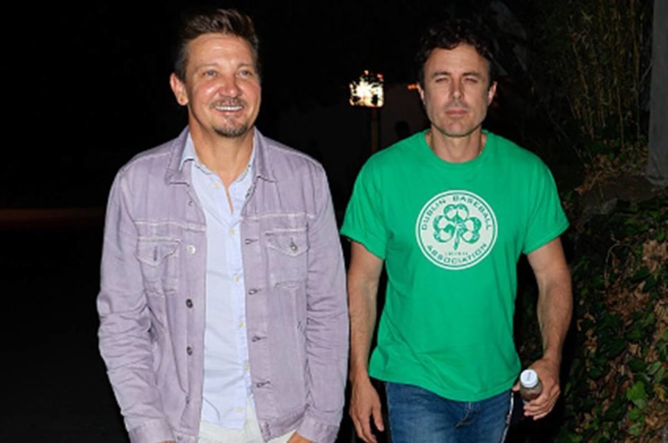 Foto colorida de Jeremy Renner usando jaqueta com amigo de blusa verde - Metrópoles