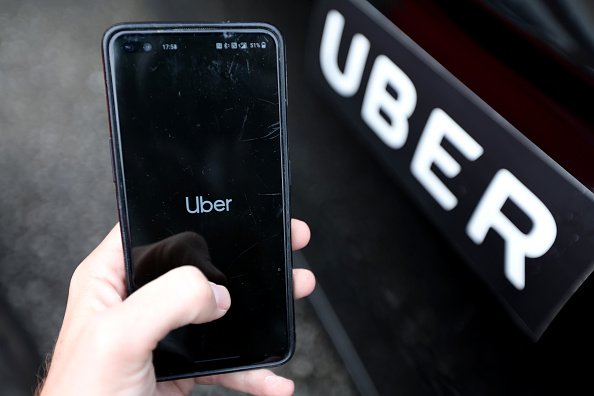 Imagem de mão segurando um celular que exibe o logotipo do Uber. Ao fundo, uma porta de carro com a marca Uber - Metrópoles